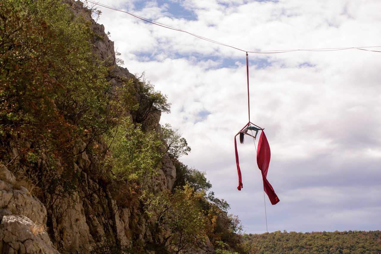 woman performing aerial silk acrobatics suspended from a red fabric high above rocky cliff and trees under cloudy sky