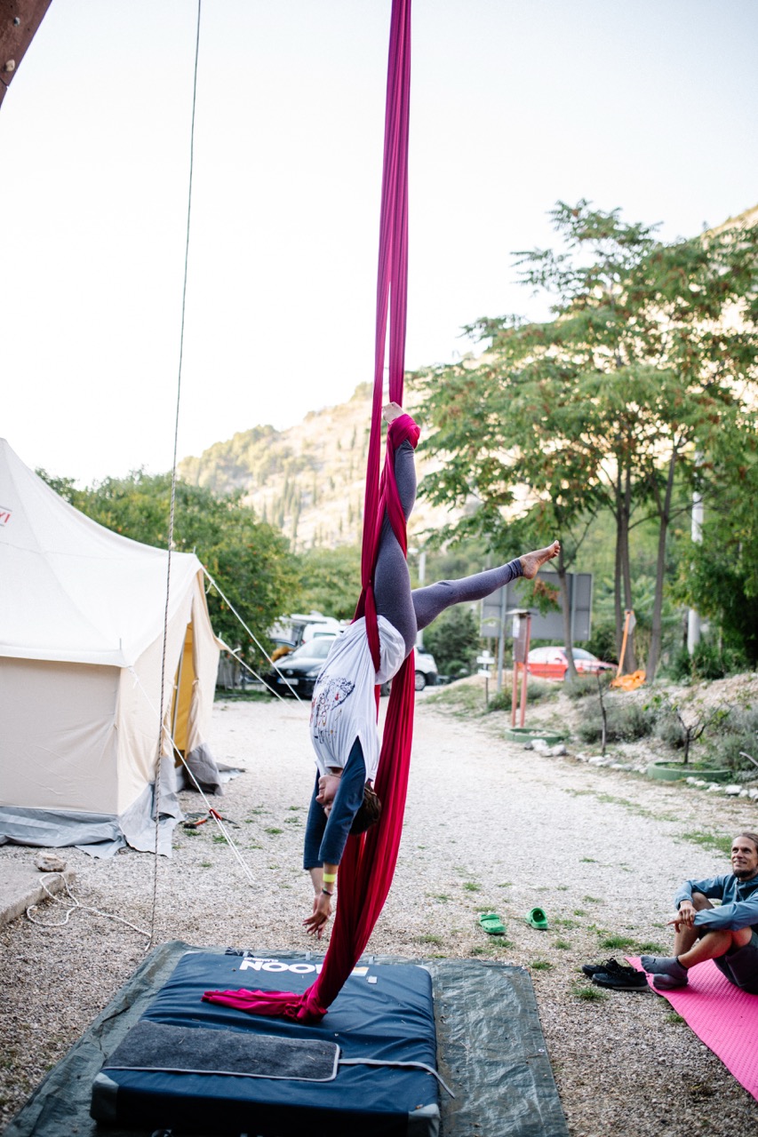 person practicing aerial silk acrobatics upside down on red fabric outdoors near a tent