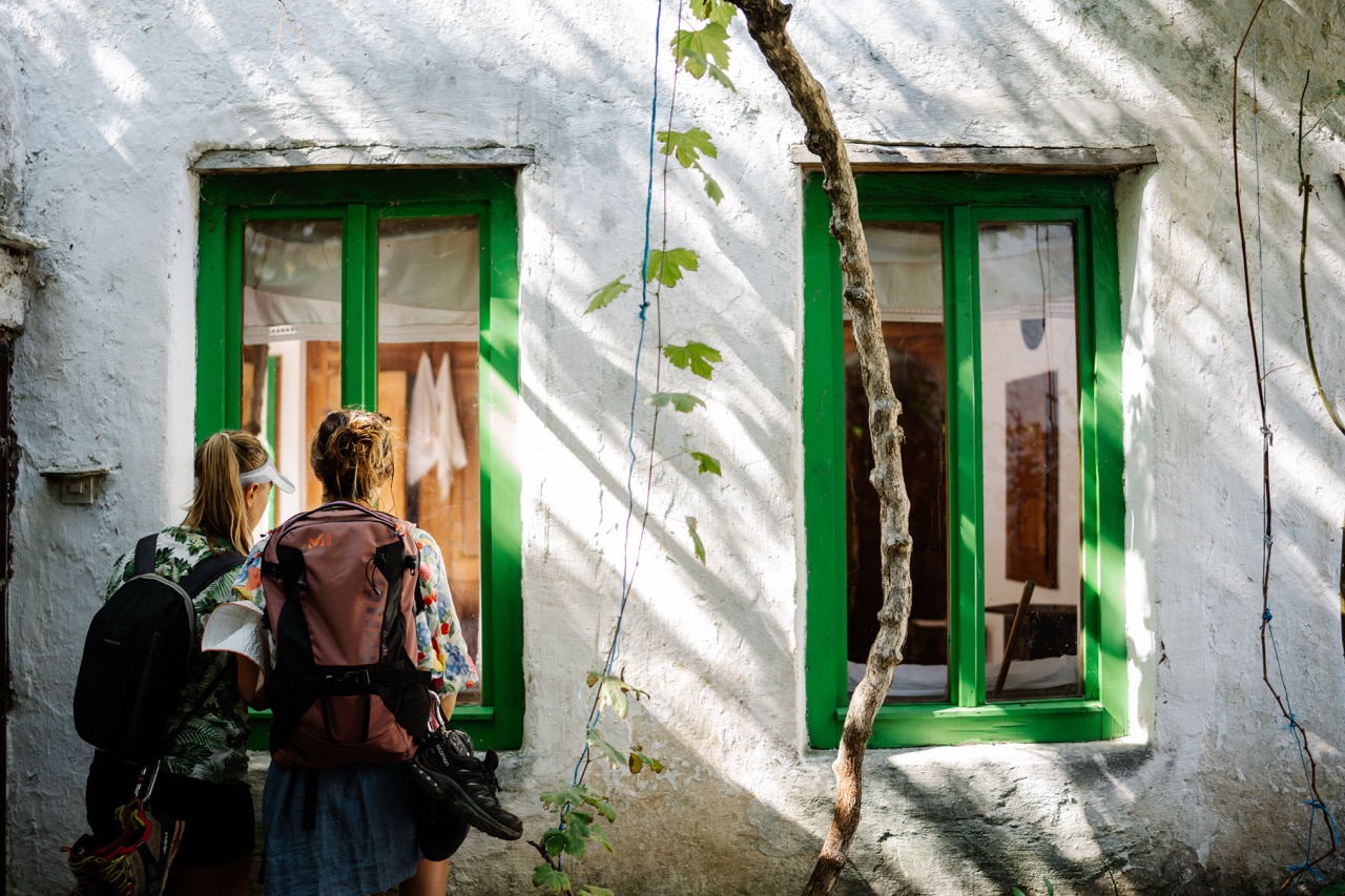 two backpackers looking through green-framed windows on a sunlit white stone wall