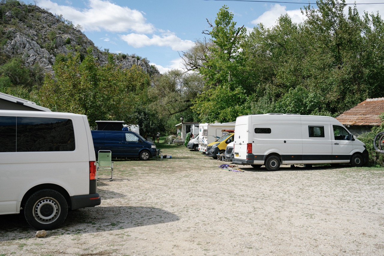 a row of camper vans and motorhomes parked on a gravel lot near green trees and rocky hills