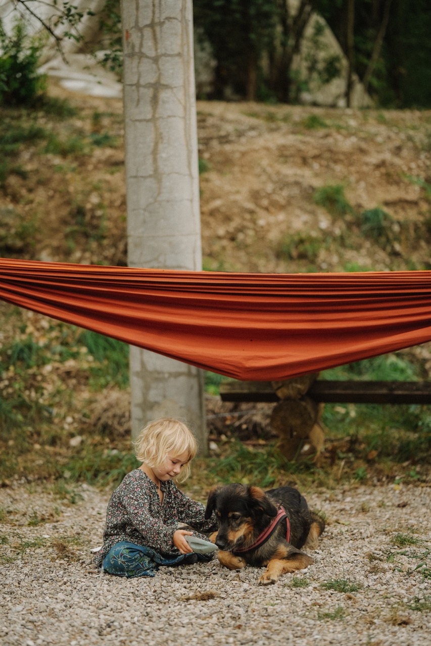 child sitting on gravel next to dog beneath a hammock in a natural setting