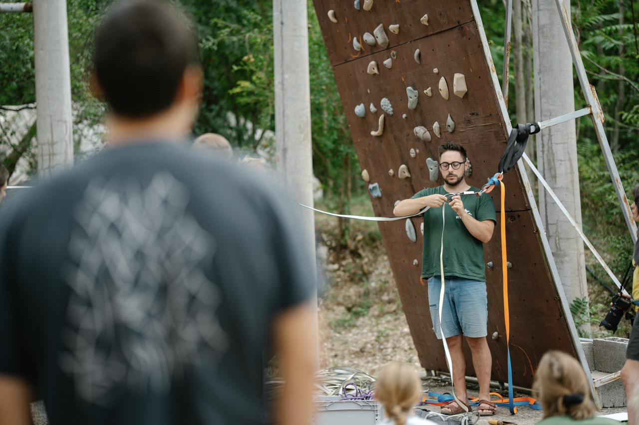 man demonstrating climbing harness and safety equipment in front of climbing wall with audience watching