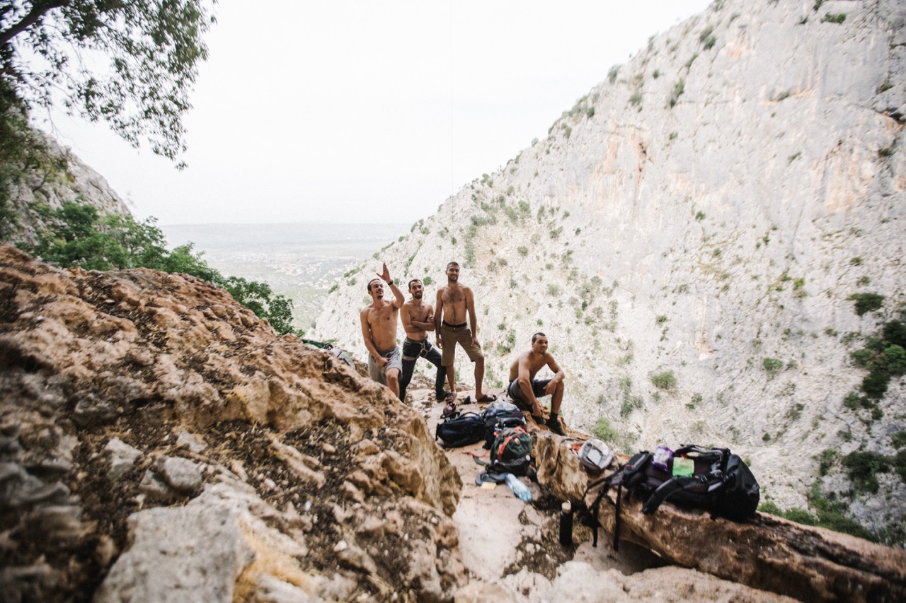 four shirtless men resting on rocky ledge with backpacks in mountainous terrain