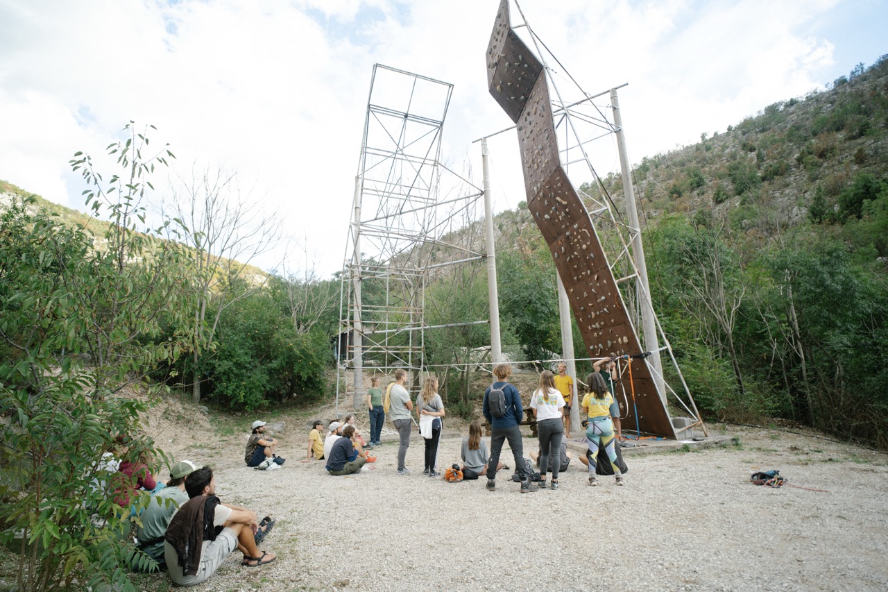 group of people gathered outdoors near a climbing wall with mountains in the background