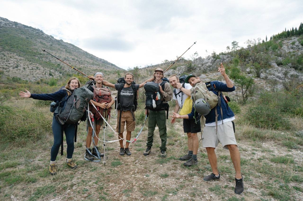 group of six hikers with backpacks posing happily on trail surrounded by hills and greenery