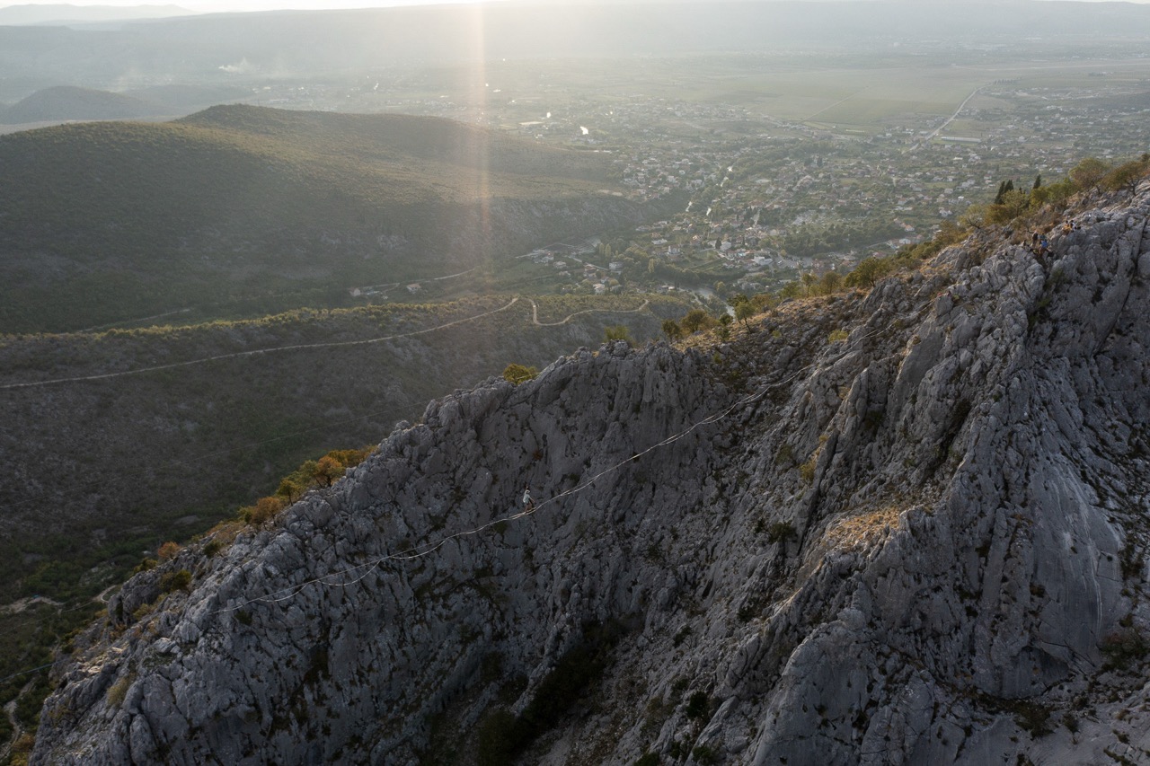 person walking on a highline across a rocky mountain ridge with a town in the valley below at sunset