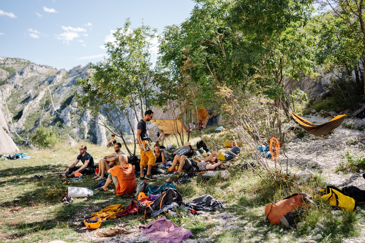 people resting and socializing under trees near mountain cliffs with camping gear around