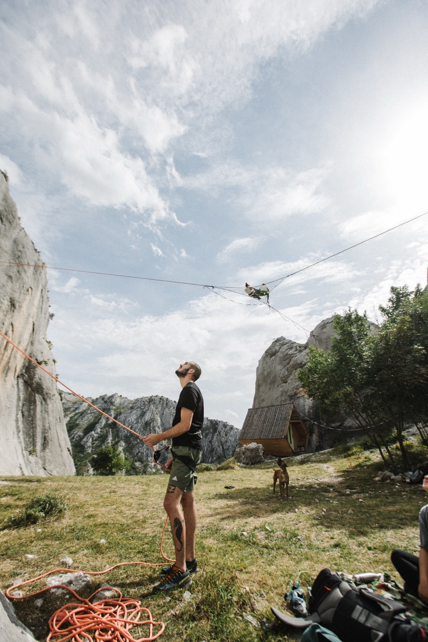 man belaying climber suspended on ropes between rocky cliffs with cabin and dog nearby