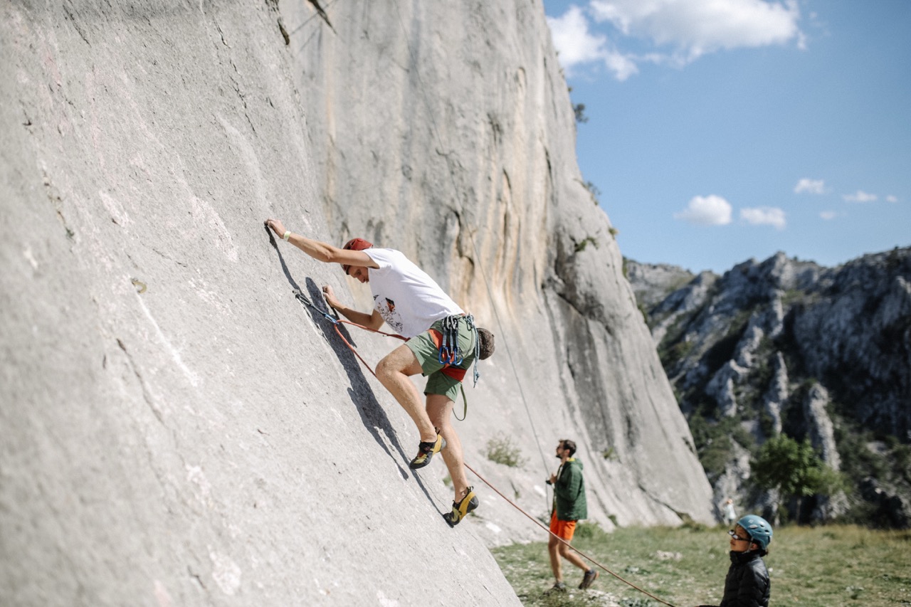 man wearing white t-shirt and green shorts rock climbing on a steep gray cliff with safety harness and rope