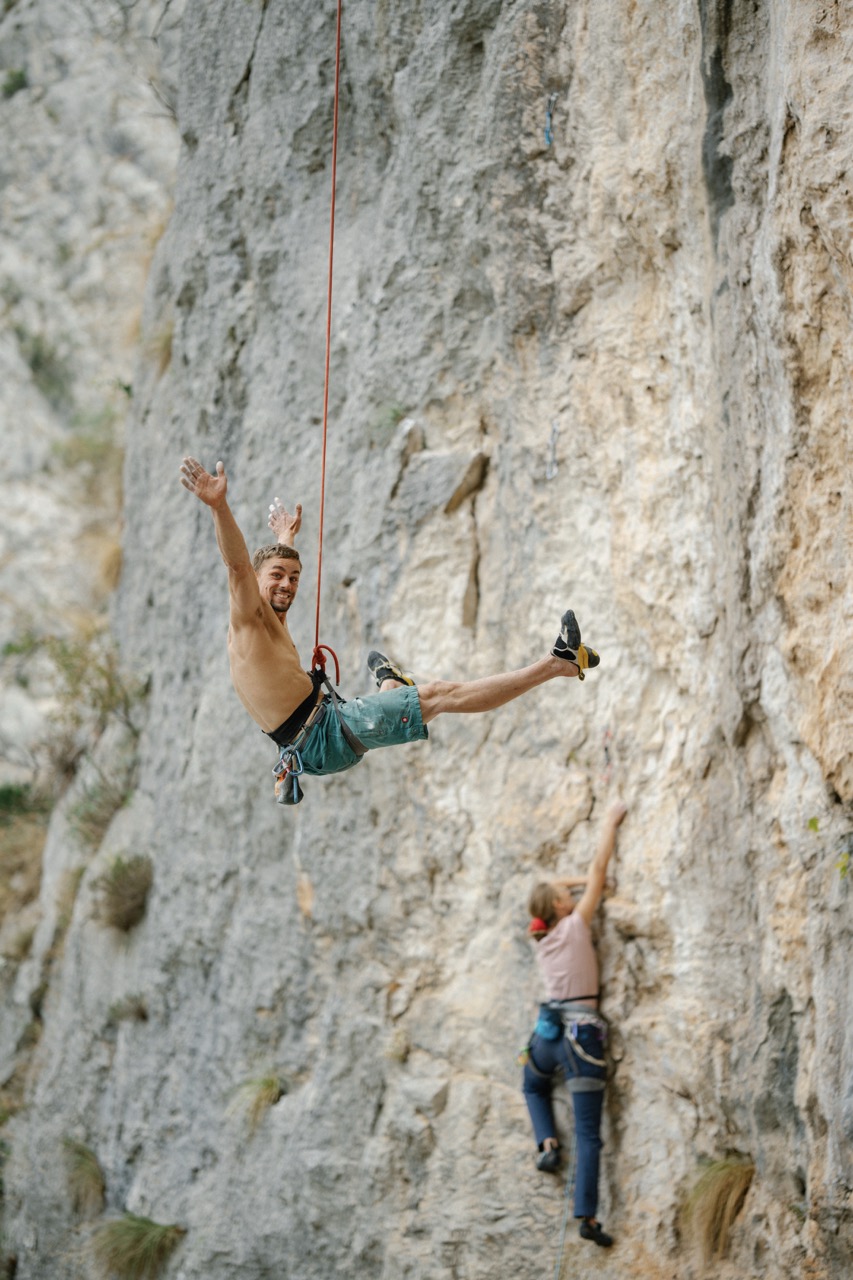 man suspended mid-air on climbing rope with woman climbing rock wall below