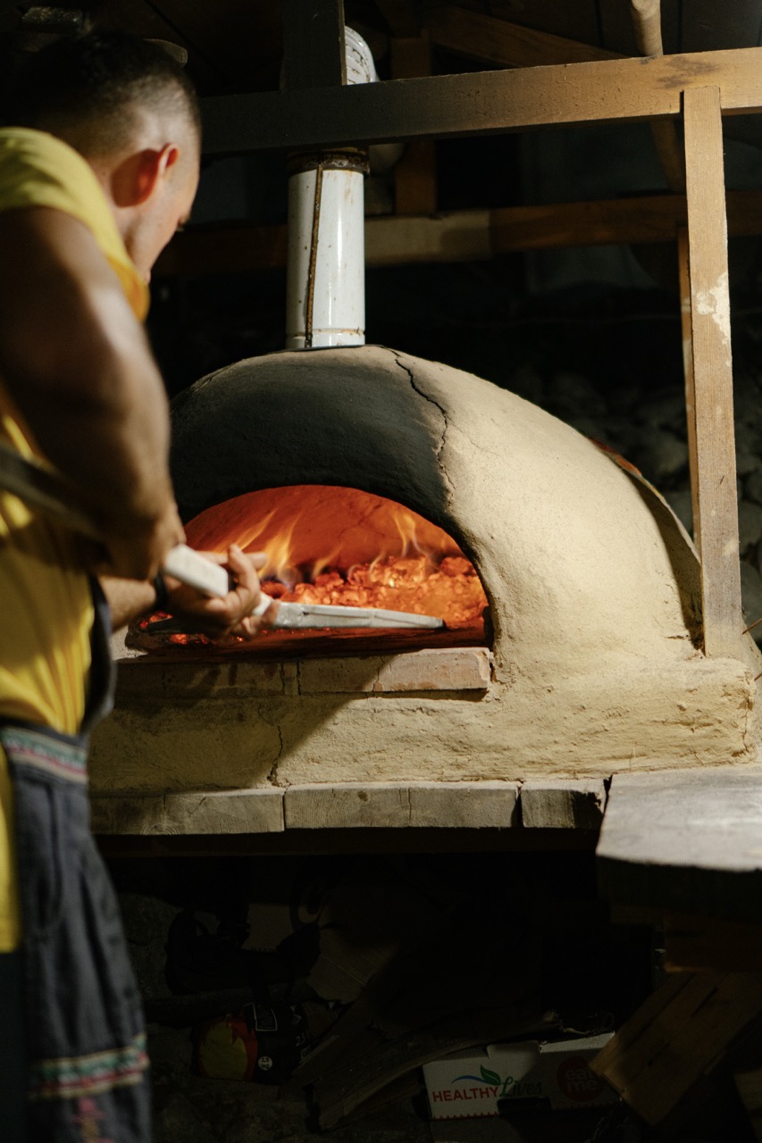 man putting pizza into wood-fired clay oven with flames inside