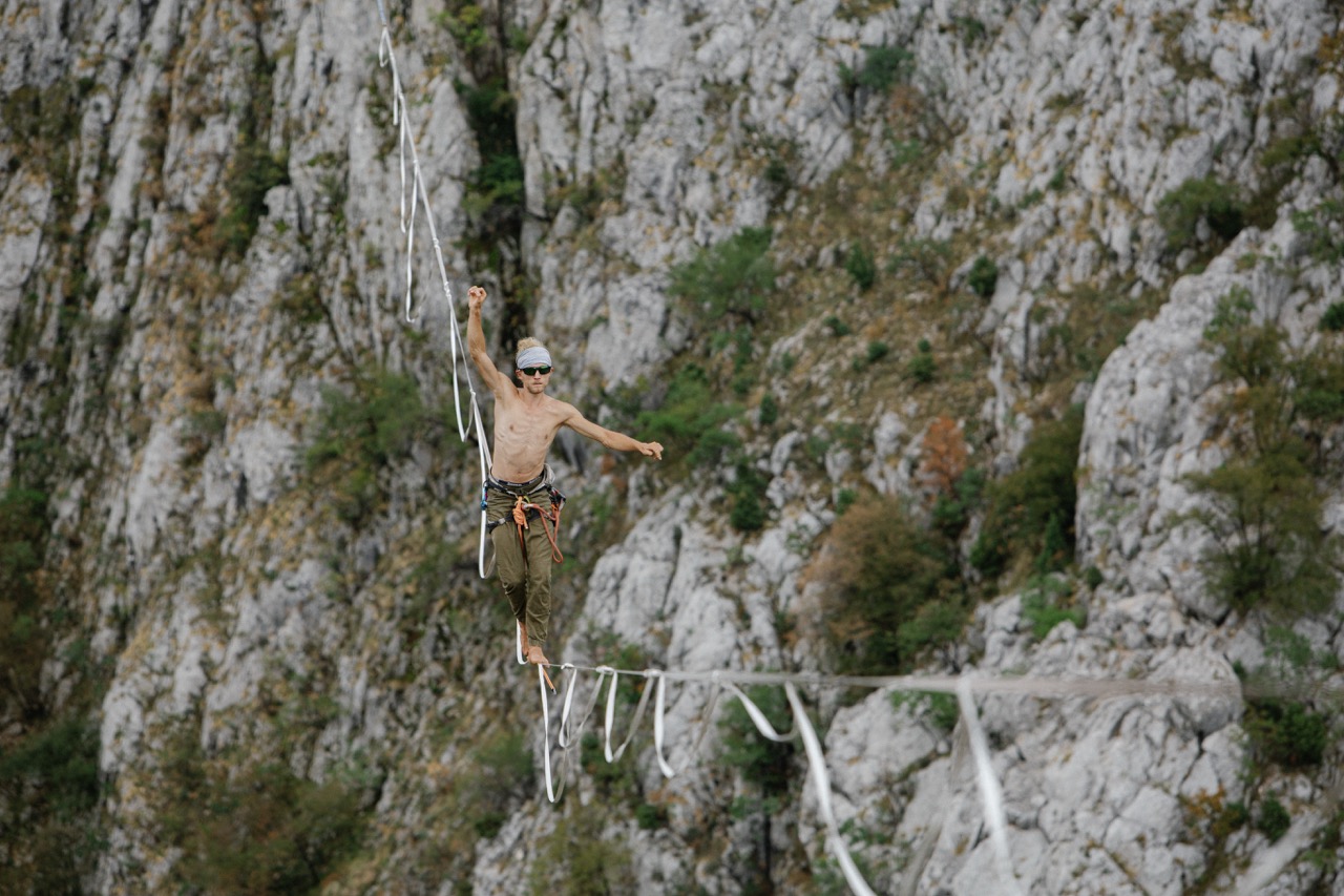man walking barefoot on a slackline stretched between cliffs wearing climbing gear and sunglasses