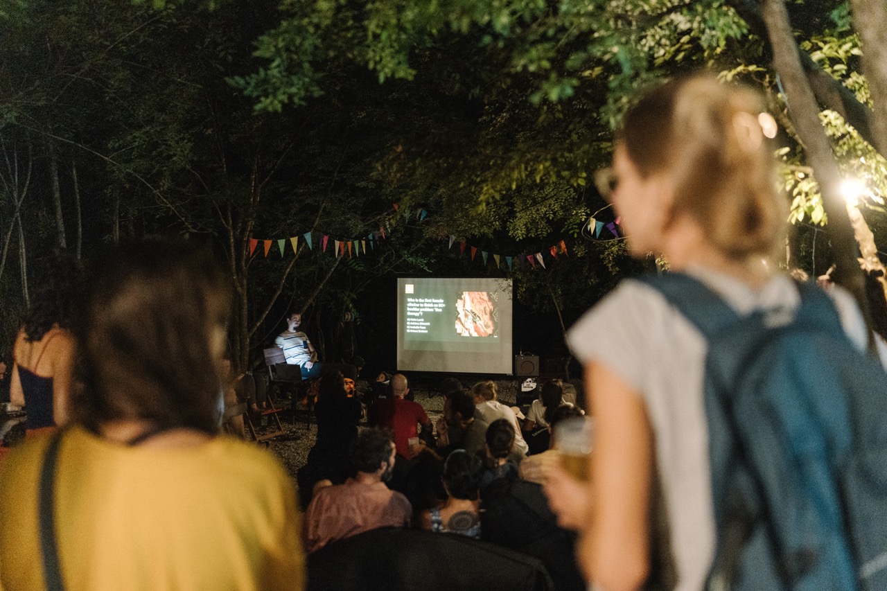 audience watching outdoor presentation on screen at night under trees
