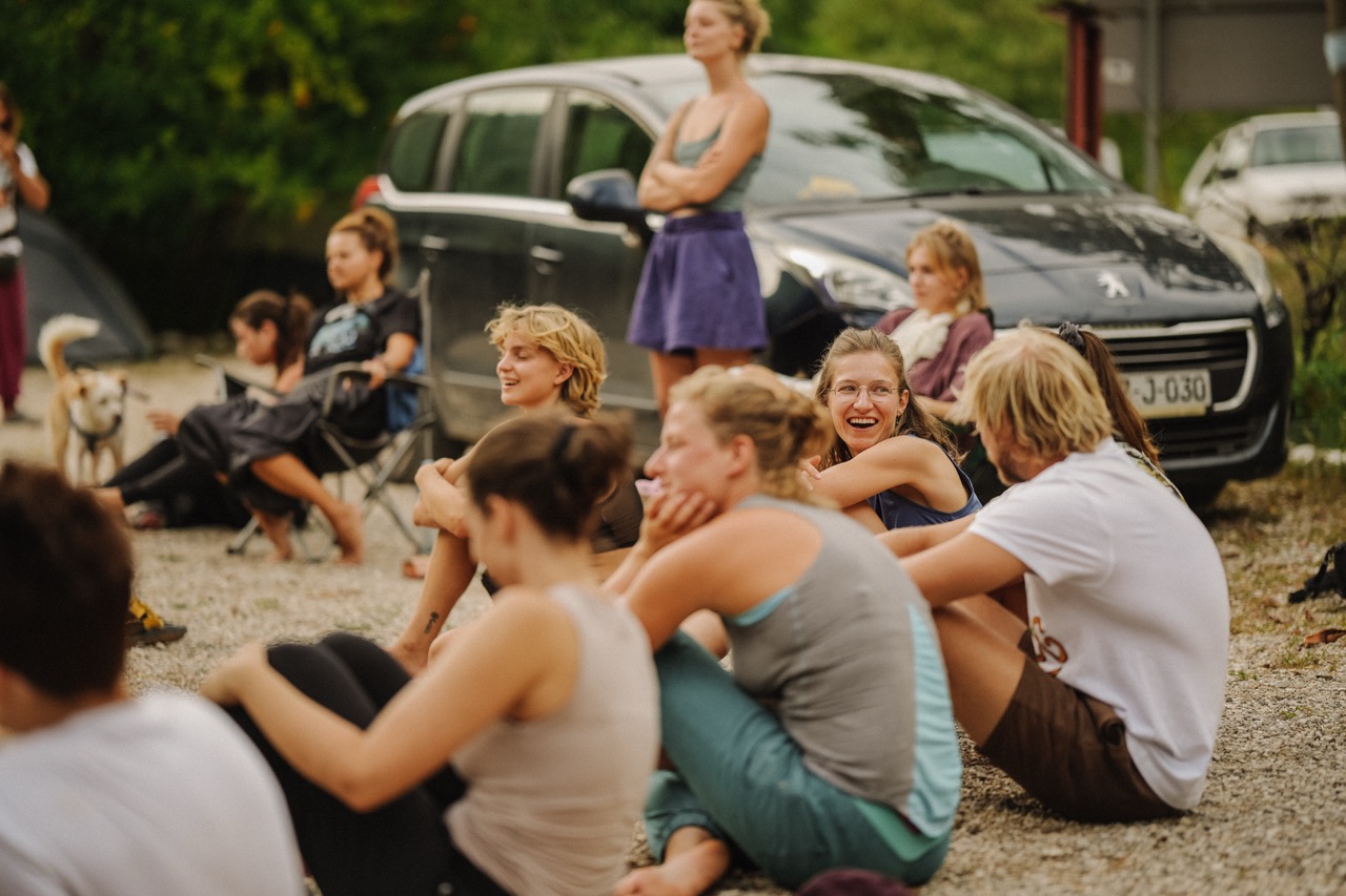 group of people sitting and standing outdoors near parked cars on a gravel surface