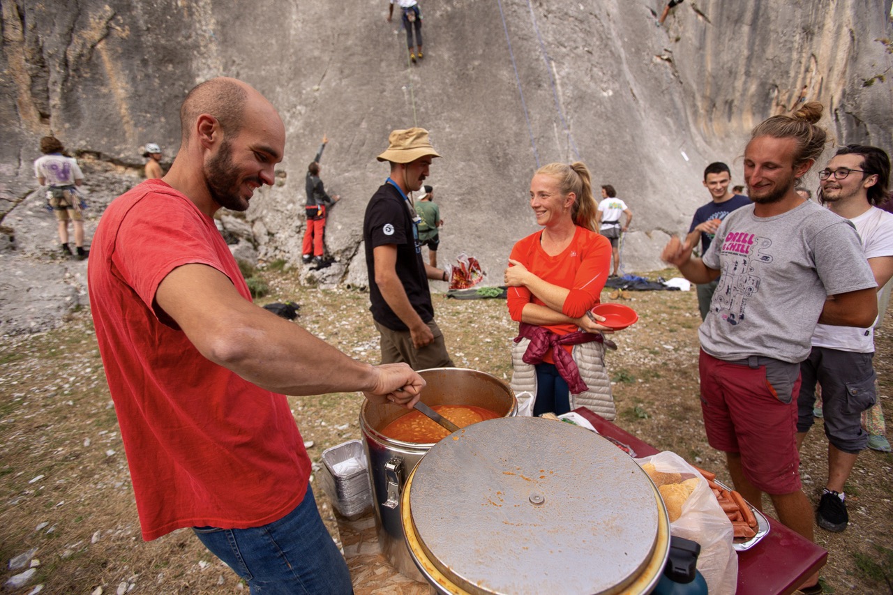 group of people enjoying outdoor meal near rock climbing area with man serving food from large pot