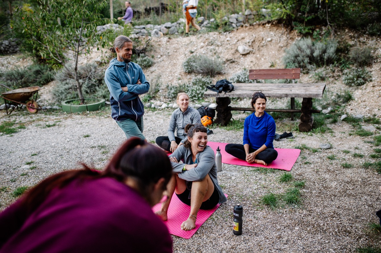 group of people sitting on pink exercise mats outdoors with one man standing nearby and trees in background