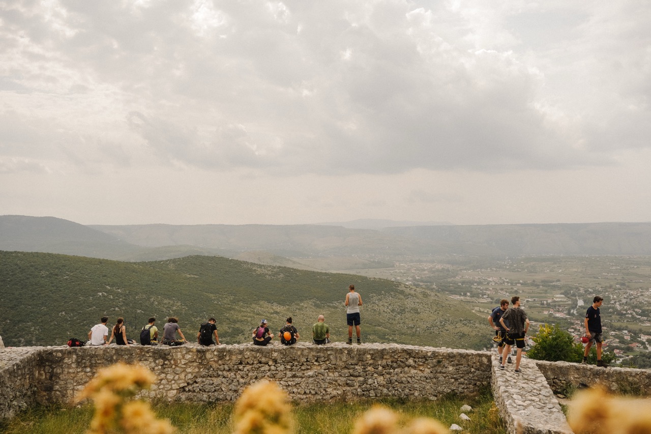 people sitting and standing on stone wall overlooking scenic green hills under cloudy sky