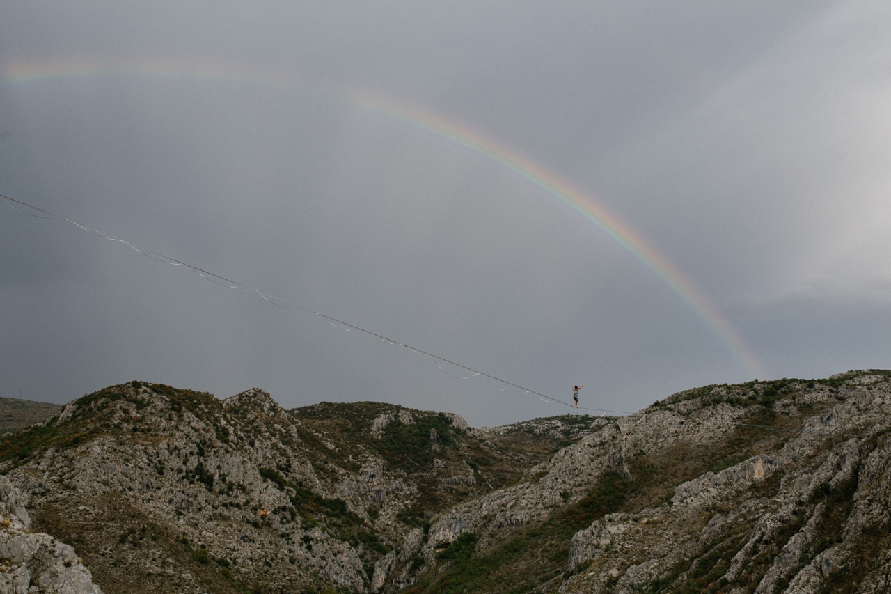 person walking on a highline between rocky hills with a faint rainbow in the background under cloudy skies