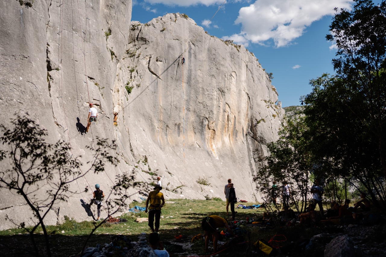 people rock climbing on large steep gray limestone cliff with trees and blue sky background