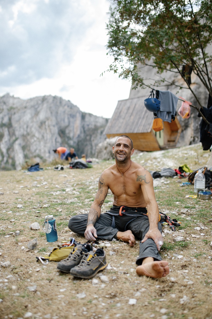 shirtless man sitting on ground near hiking boots and outdoor gear with rocky mountains and wooden cabin in background
