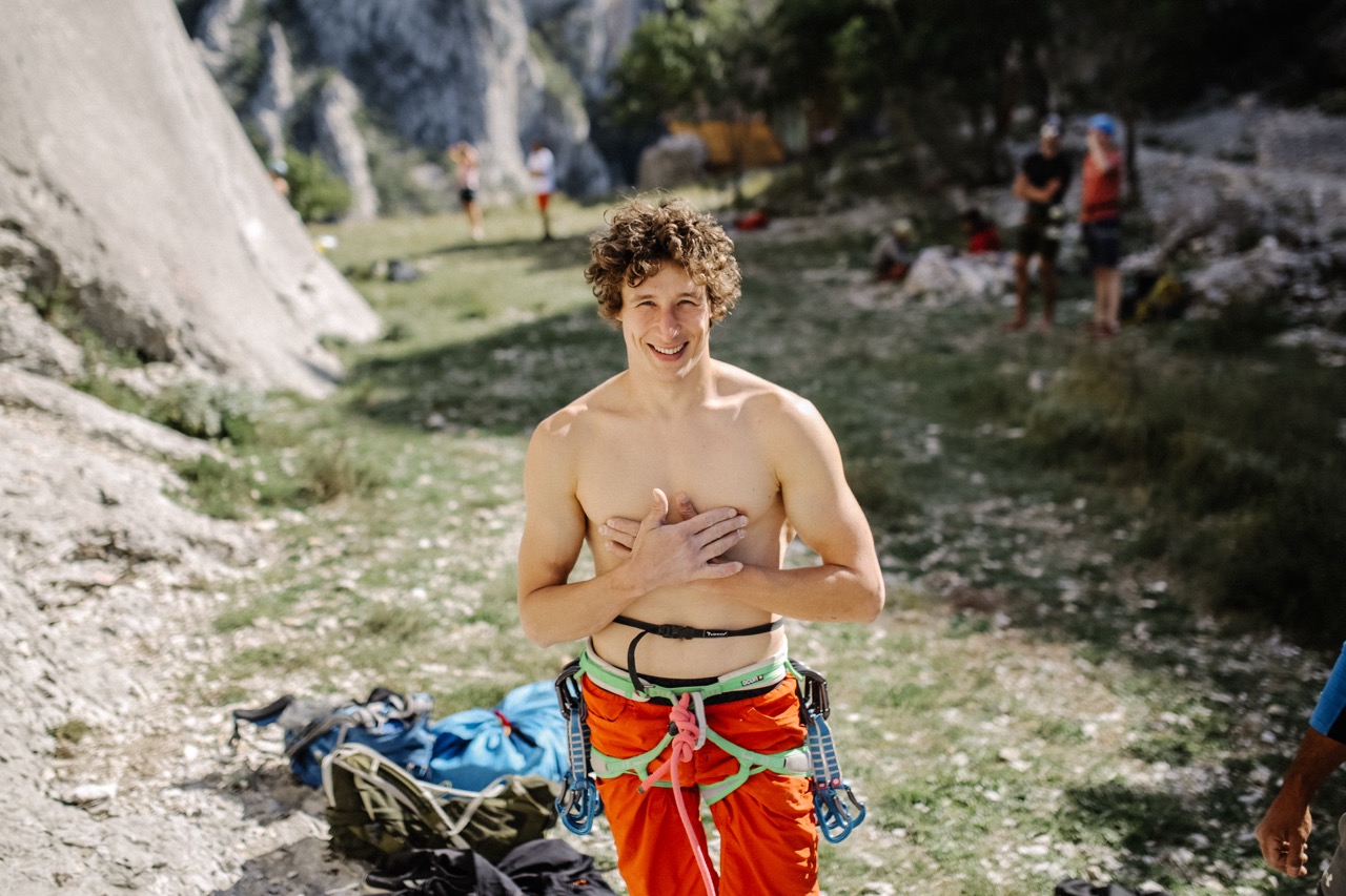 smiling shirtless man in orange climbing pants wearing a harness outdoors