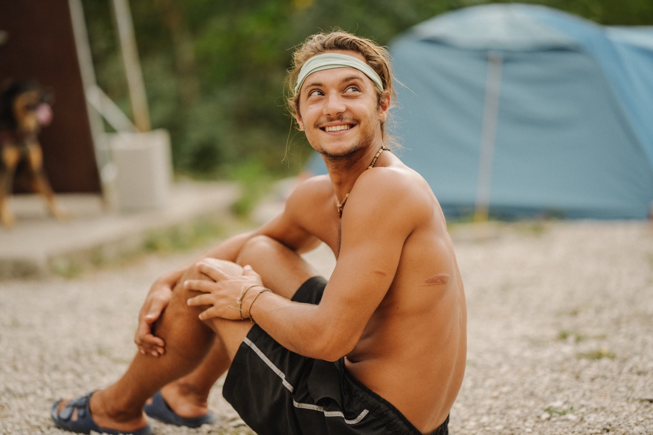smiling shirtless man sitting on ground near a blue tent and looking to the side