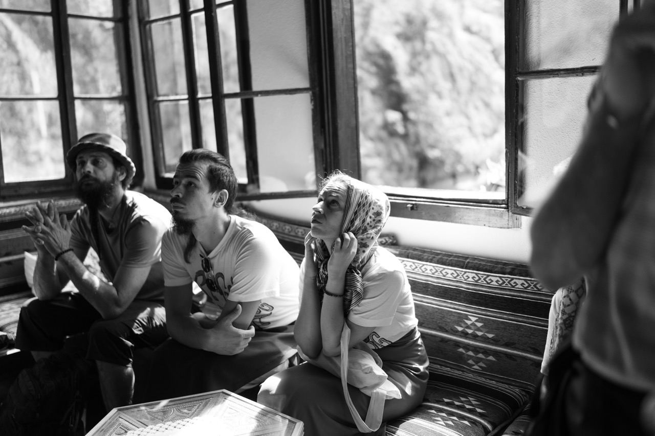 three people sitting closely on a patterned bench near open windows looking attentively upwards in black and white