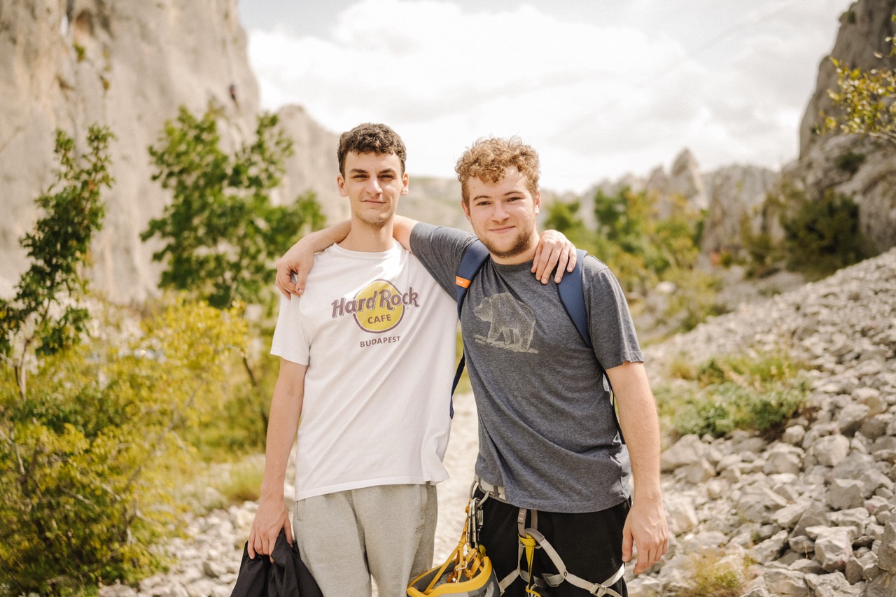 two young men standing on rocky trail with climbing gear and backpack in mountainous landscape