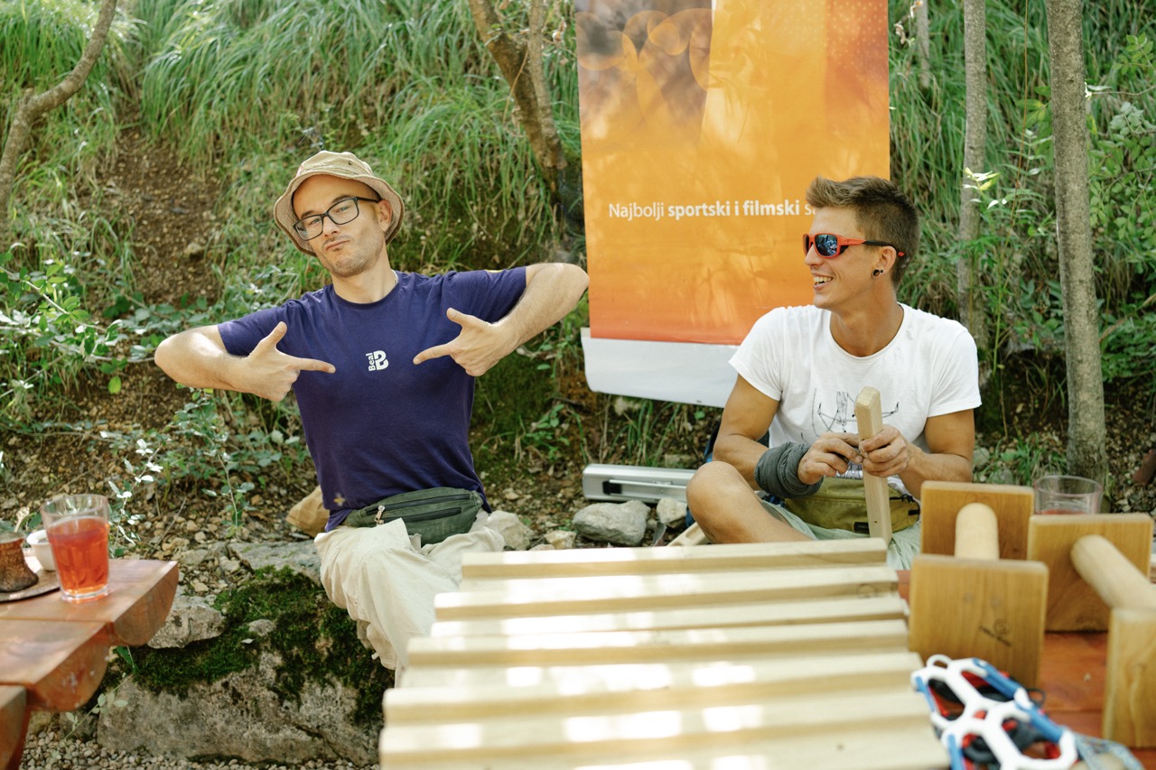 two young men sitting outside in a green area, one pointing to his navy t-shirt and the other smiling with sunglasses
