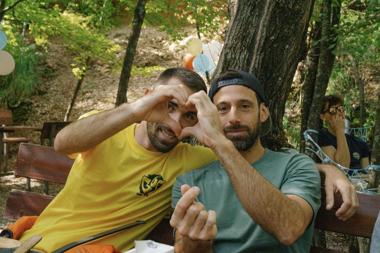 two men sitting on a bench making heart and finger heart hand gestures outdoors