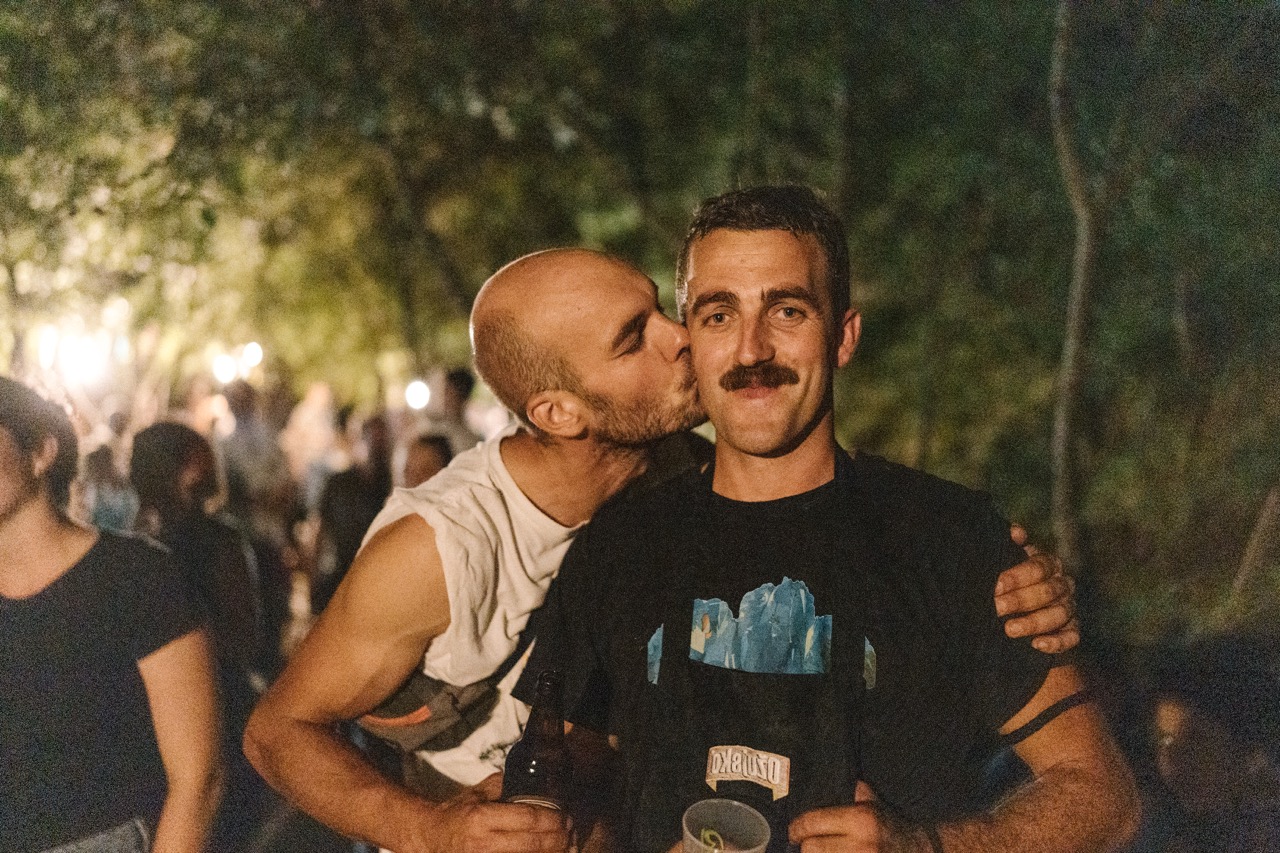 two men at an outdoor night party with one kissing the other on the cheek