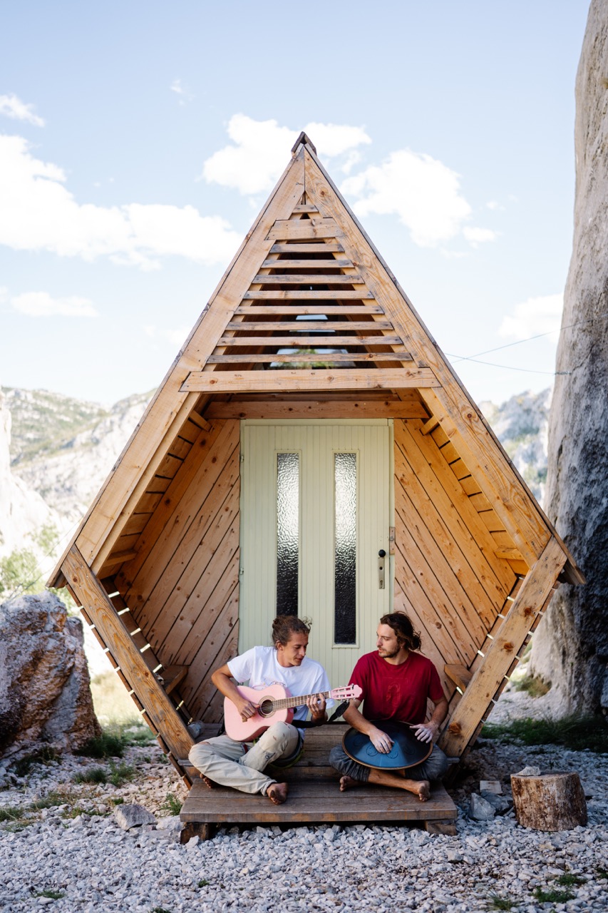 two young men playing a pink guitar and a handpan while sitting barefoot on the wooden porch of an angular wooden cabin in a rocky natural setting