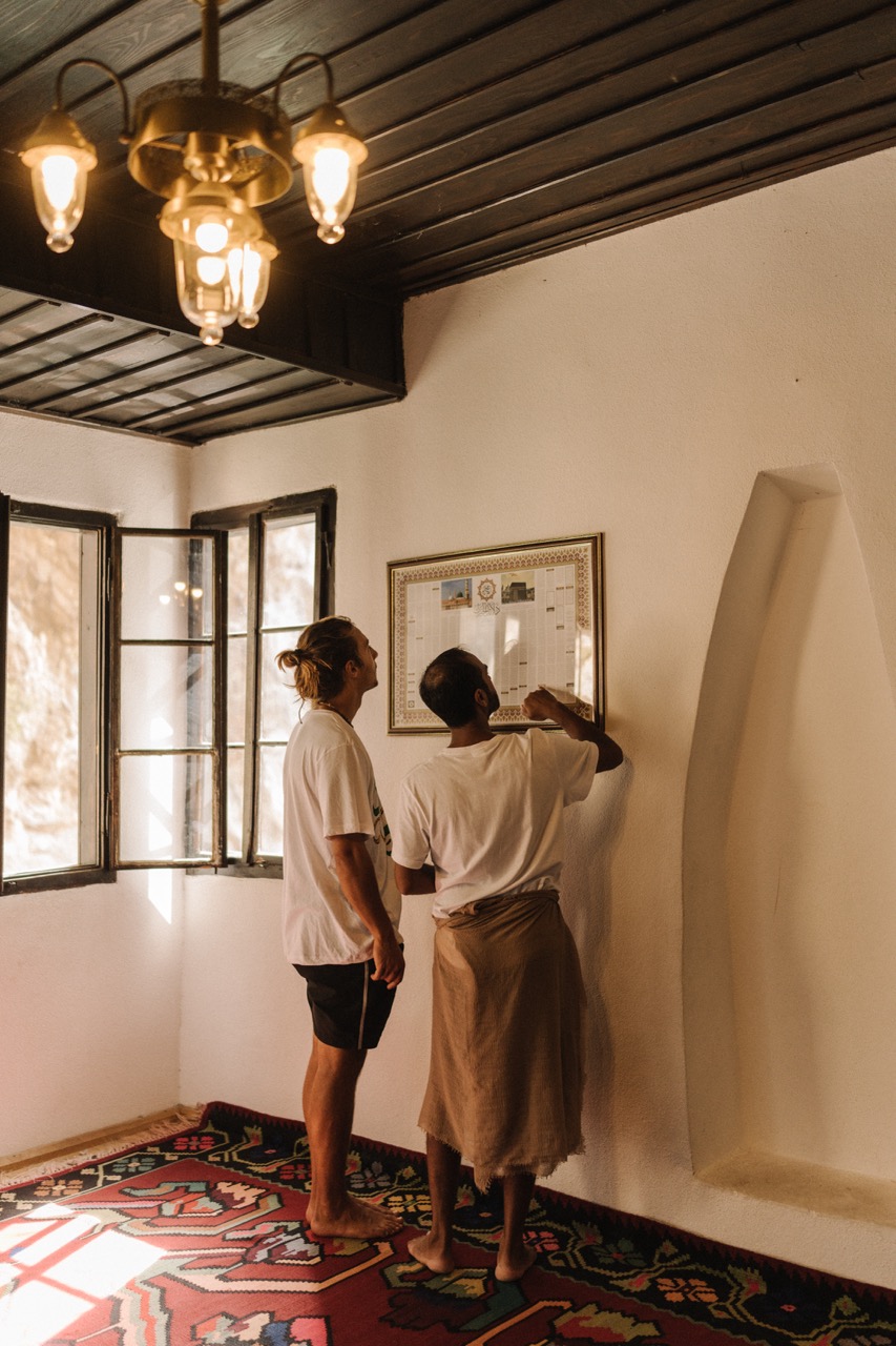 two men reading framed artwork on white plaster wall in a cozy room with wooden ceiling and colorful carpet