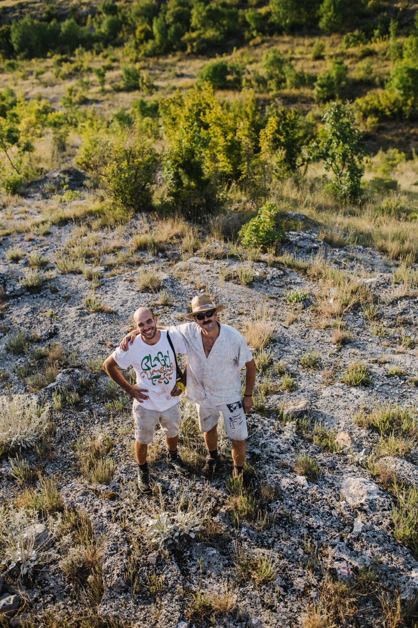 two men standing on rocky terrain with green bushes in the background during daylight