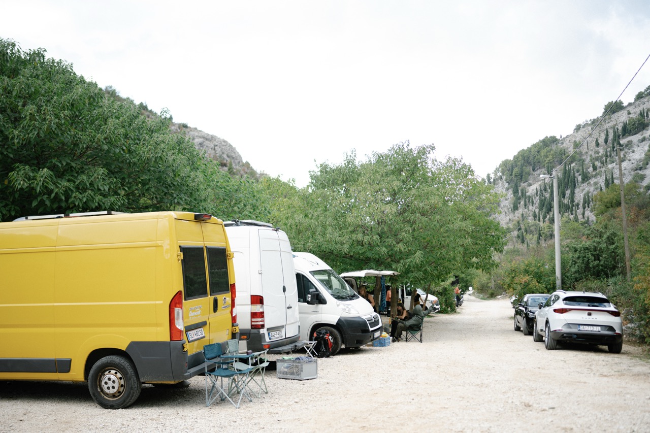 yellow and white vans parked on a dirt road with people sitting under green trees in a mountainous area