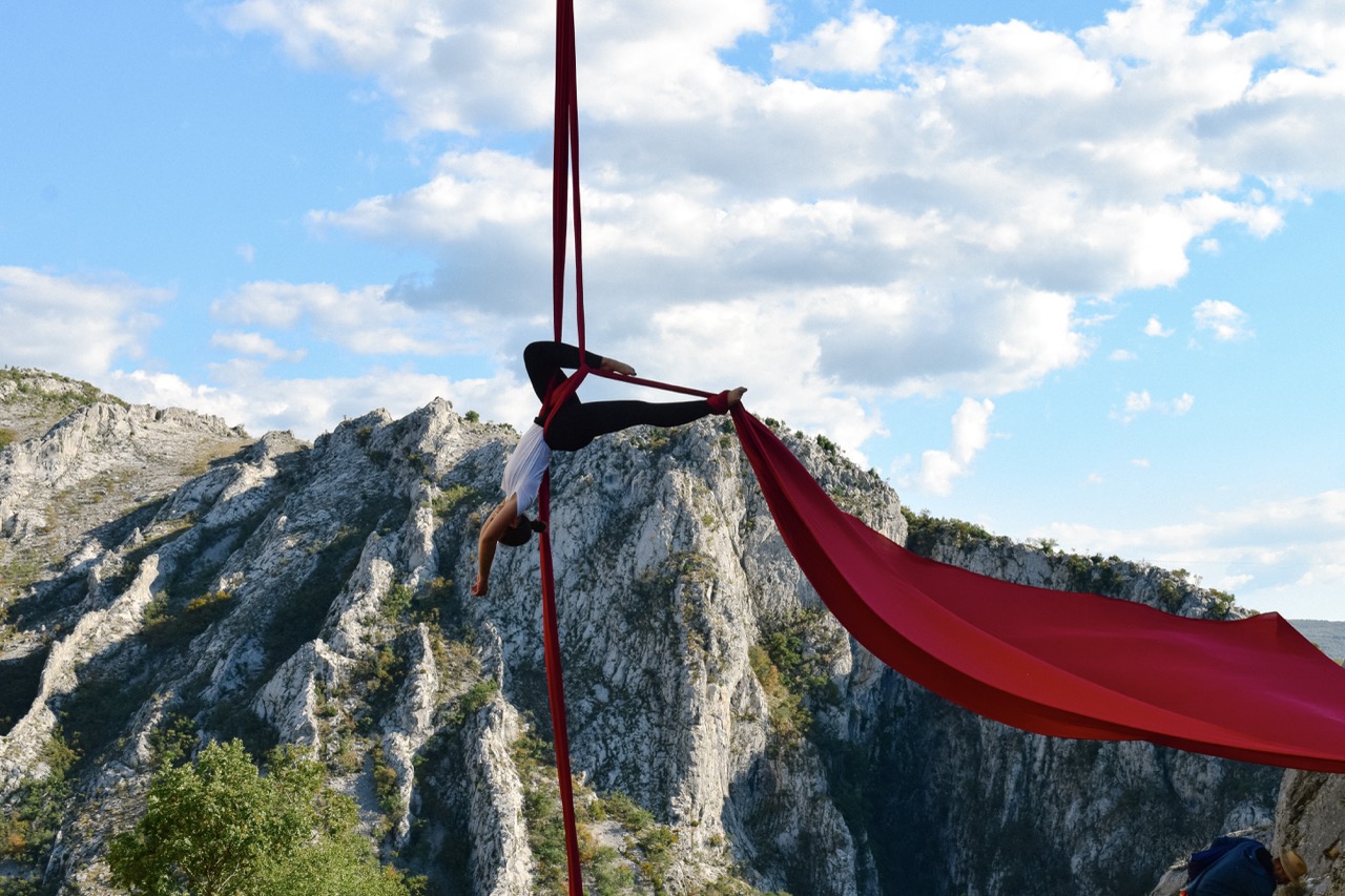 woman performing aerial silk acrobatics outdoors with red fabric against mountain landscape