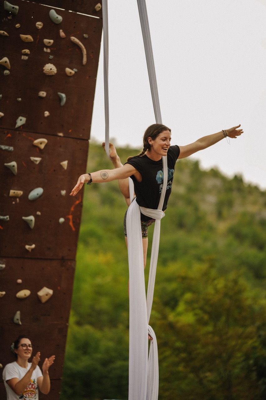 woman performing aerial silk act outdoors near a rock climbing wall while another person claps