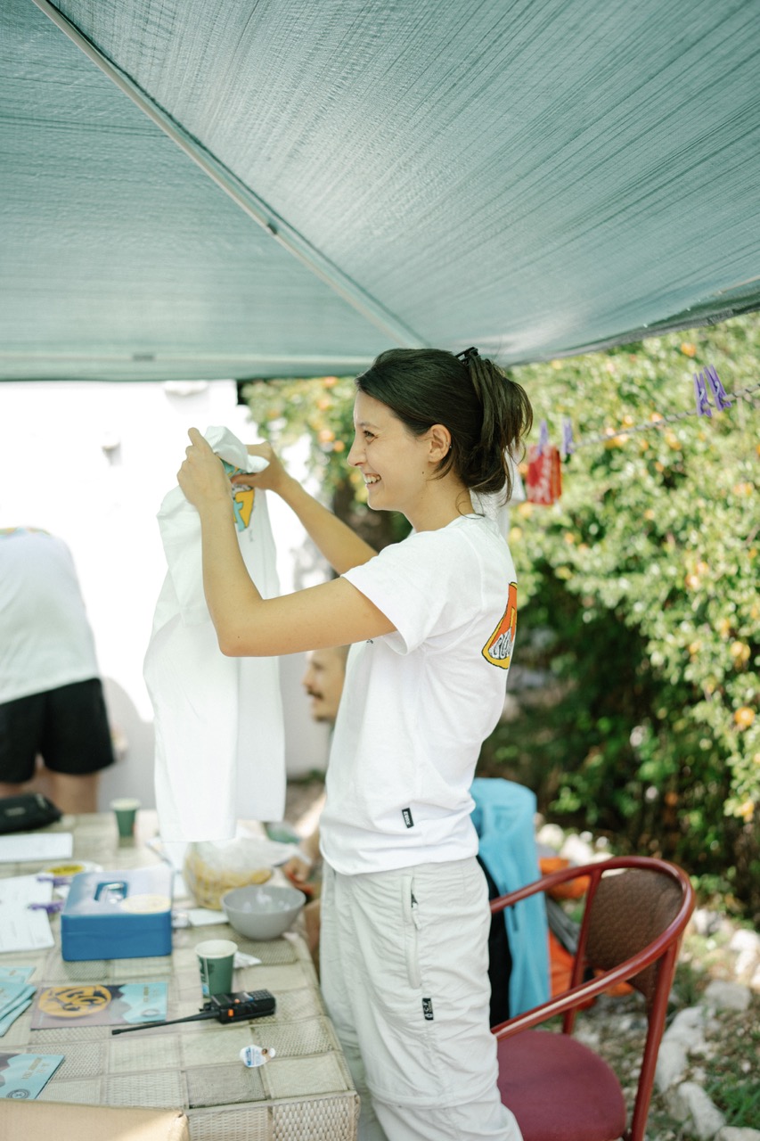 woman folding a white t-shirt with a colorful logo at an outdoor table under a canopy